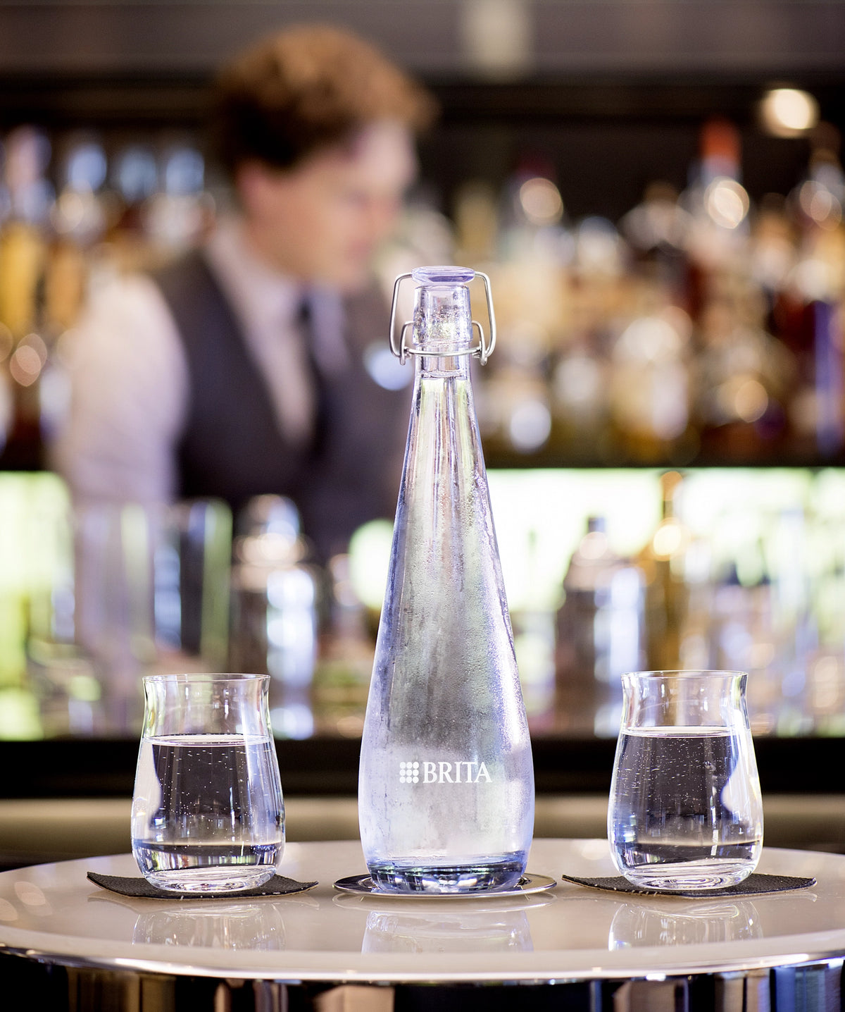 BRITA Bottiglia Swing con due bicchieri d'acqua su un bancone da bar, con un barista sfocato sullo sfondo.