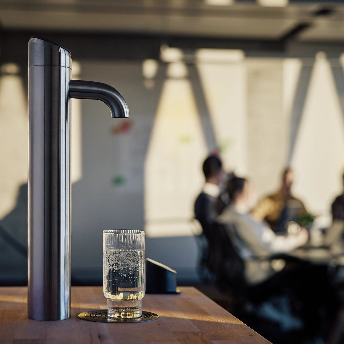 Distributore d'acqua BRITA Extra I-Tap su un tavolo di legno, con un bicchiere d'acqua sotto, in un ambiente d'ufficio sfocato.
