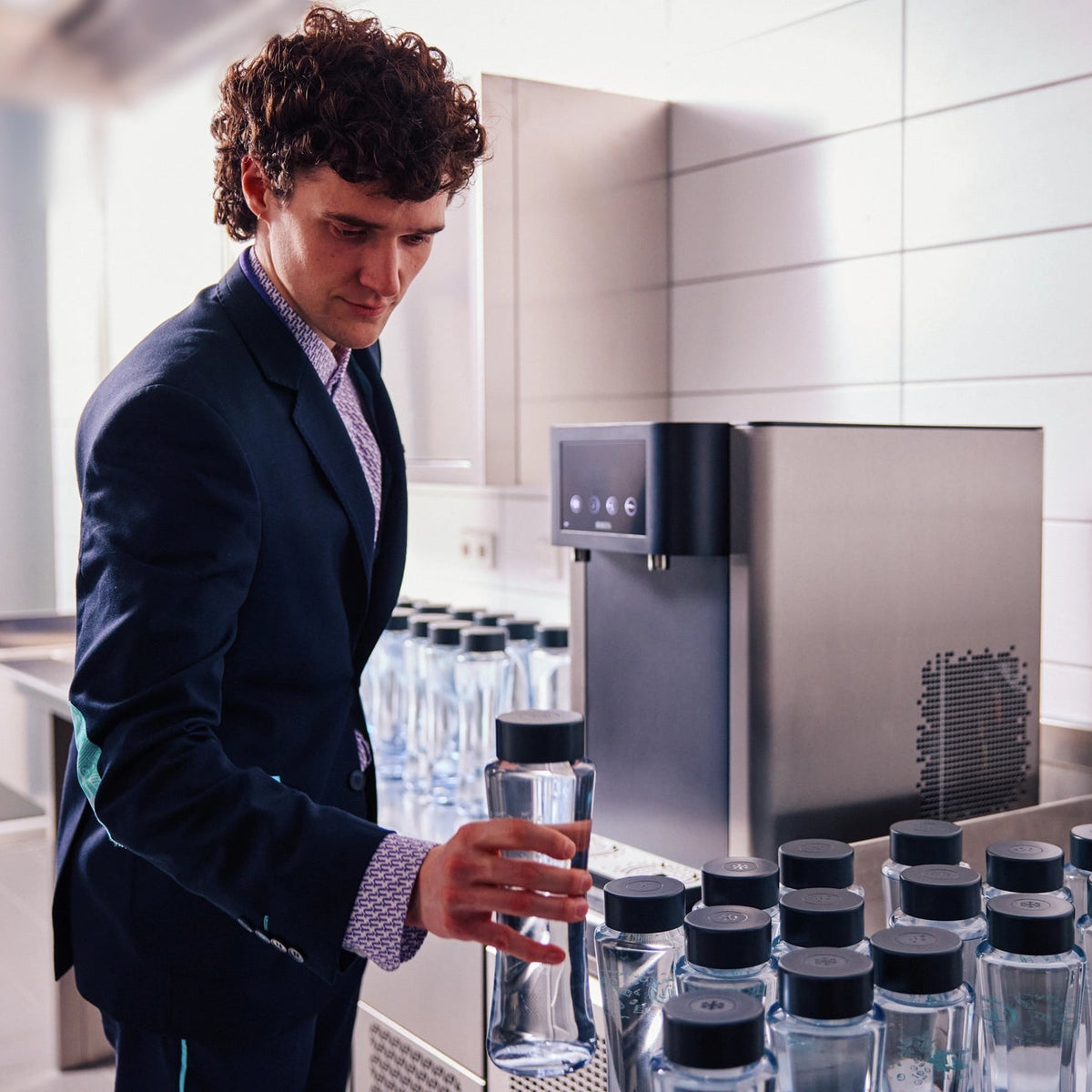 Un uomo in giacca e cravatta in piedi davanti a un distributore d'acqua BRITA Fill Pro con diverse bottiglie Twist.