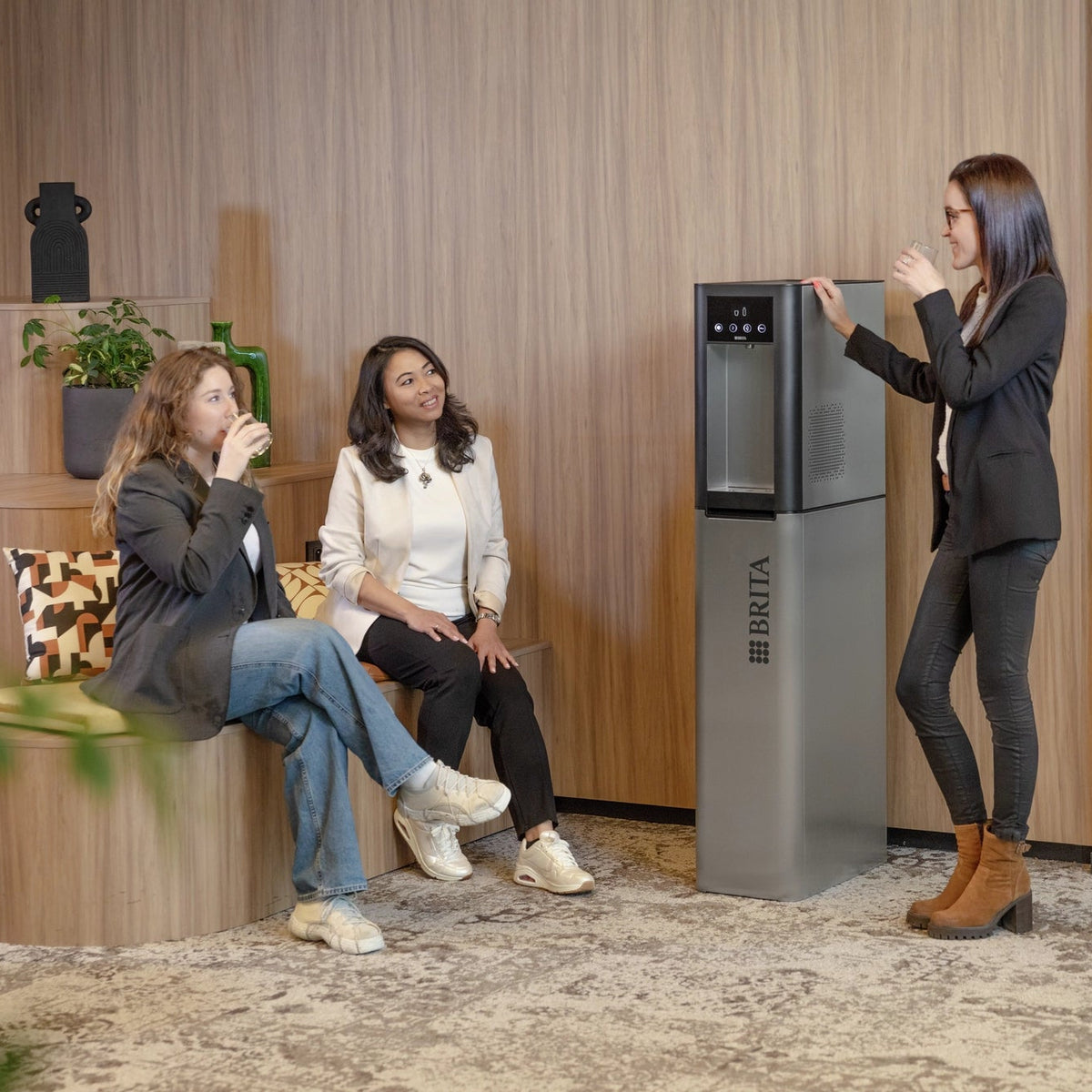 Donna in piedi accanto a un distributore d'acqua Top Pro in un ambiente d'ufficio moderno, con altre due donne sedute su una panchina.
